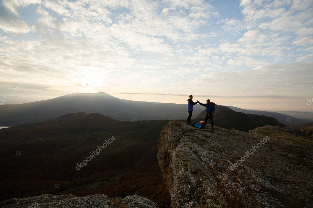 Two climbers standing on summit above clouds in the mountains holding hands. Silhouettes of hikers celebrating ascent on sunset. Help, cooperation, synergy, friendship concept.