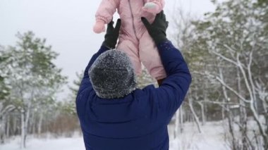 Baba, güzel kızını karlı kış gününde havaya fırlatıyor. Ebeveyn çocuğunu yukarı fırlatır ve parkta yakalar. Pembe tulumlu şirin bebek gülüyor ve gülümsüyor. Çocuklar kış modası.