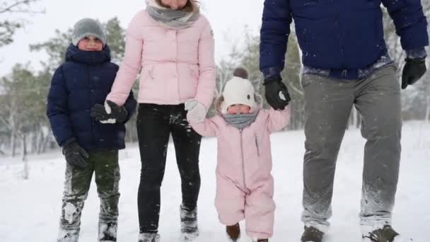 A baba meleg kezeslábasban csinálja az első lépéseket. A gyerek megtanul járni. Barátságos teljes család séta téli erdőben nyaralni hóesés alatt. Apa, anya, fia és lánya karácsonykor a szabadban — Stock videók