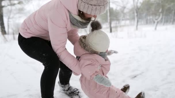 Mutter zieht Baby aus dem Schnee. Kind im warmen Overall lernt laufen. Mädchen, das im Winterwald bei Schneefall die ersten Schritte macht. Frau mit Tochter an einem verschneiten Tag im Freien. warmer rosa Strampler. — Stockvideo