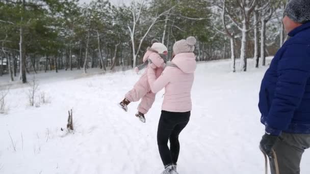 Lycklig kvinna höjer och snurrar med barnet i varm overall i händerna på snöig dag. Mamma har kul med dottern i slow motion. Söt familj promenader i vinterskogen på semester under snöfall. — Stockvideo