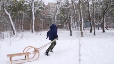 Sıcak giysili yakışıklı çocuk, karlı kış gününde tahta kızak çekiyor. Küçük adam kar yağarken parkta kayıyor. Donanma ceketli bir çocuğun ormanda kızakla kaymak için tepeye tırmanırken görüntüsü. Yavaş çekim.