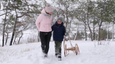 Sıcak giysili tatlı bir çocuk, karlı kış gününde annesiyle ahşap mahsul kızağı çekiyor. Küçük adam kar yağarken parkta kayıyor. Ormanda kızakla kaymak için tepeye tırmanan çocuklu bir kadın..