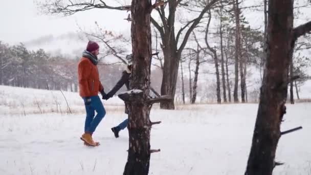 Young loving couple walking in a park on snowfall. Man following his girlfriend holding hands, enjoying snow on winter day in snowy forest. Hipster woman in stylish clothing leads boyfriend by hand. — Stockvideo