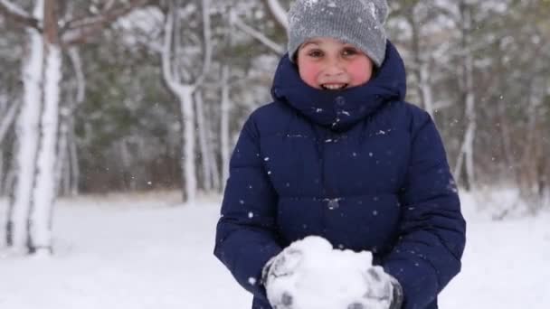 Szczęśliwy chłopak w marynarce bawi się śniegiem i wyrzuca go w powietrze w lesie w mroźny zimowy dzień. Wesołe dziecko w stylowym zimowym stroju cieszą się pierwszą śnieżną pogodą w parku leśnym. Zwolniony ruch. — Wideo stockowe