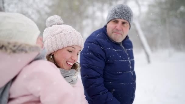 Amistosa familia completa caminando a través del bosque de invierno de vacaciones bajo las nevadas. Padre, madre e hija descansan en las vacaciones de Navidad al aire libre en el día nevado. Bebé en mameluco rosa . — Vídeos de Stock