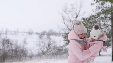 Mutlu kadın, karlı bir günde bebeği sıcak tulumla kucaklar ve kucaklar. Gülümseyen anne kızıyla yavaş çekimde eğleniyor. Kış ormanlarında kar altında gezen sevimli bir aile..