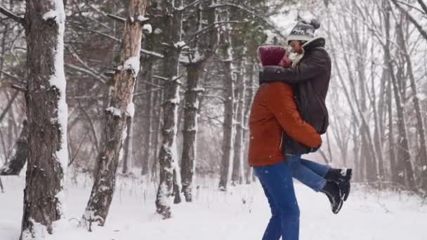 Ο άνθρωπος γυρίζει ή γυρίζει ευτυχισμένη φίλη της κρατώντας της στα χέρια του σε χιονόπτωση σε πευκοδάσος. Αγαπημένο ζευγάρι διασκεδάστε στο χιονισμένο χειμερινό πάρκο. Ημέρα του Αγίου Βαλεντίνου και Χριστούγεννα. Γυναίκα γελάει σε αργή κίνηση. — Αρχείο Βίντεο