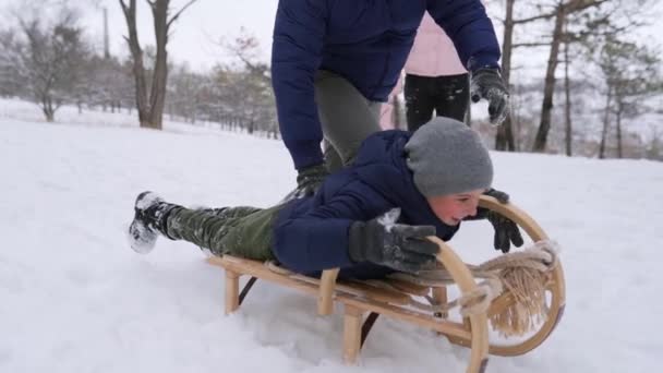 Apa elindítja fiát, amint szánkót nyom le a hegyről egy havas téli napon a parkban. Fiú szánkó a szabadban. Boldog családi szánkózást. Az emberek szánkóznak és élvezik a karácsonyi vakációt az erdőben. Lassú mozgás.. — Stock videók