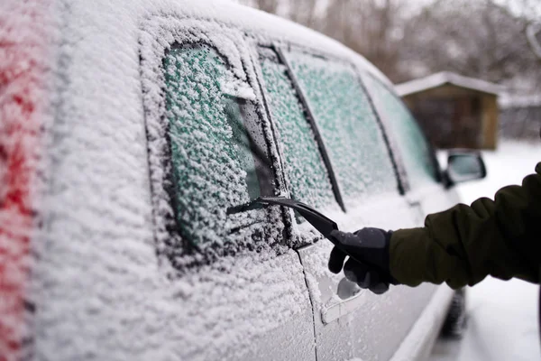 Cleaning the side car windows of snow with ice scraper before the trip. Man removes ice from car windows. Male hand cleans car with special tool at snowy frosty winter day.