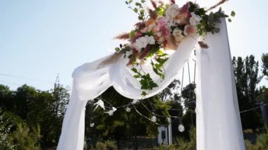 Düğün kemeri, güneşli yaz gününde çiçeklerle süslenmiş. Şenlikli çiçek süslemesi. Güller ve tüy çimleri beyaz tekstil çerçevesinde ampul çelengi ile.