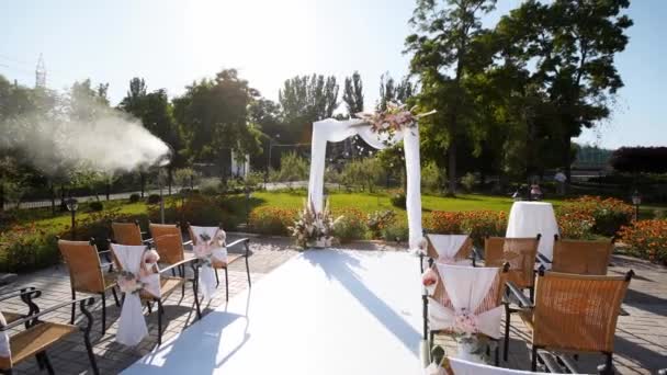 在阳光明媚的夏日,婚礼拱门装饰着鲜花. 椅子上的节日花卉装饰。 白色纺织品框架上有灯泡花环的玫瑰和羽草. — 图库视频影像