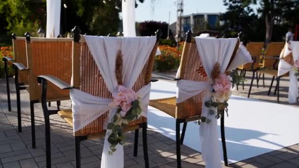 Lugar de ceremonia de la boda decorado con flores en el soleado día de verano. Decoración floral festiva en sillas. Rosas y hierba de plumas con guirnalda bulbo sobre arco textil blanco sobre fondo . — Vídeos de Stock