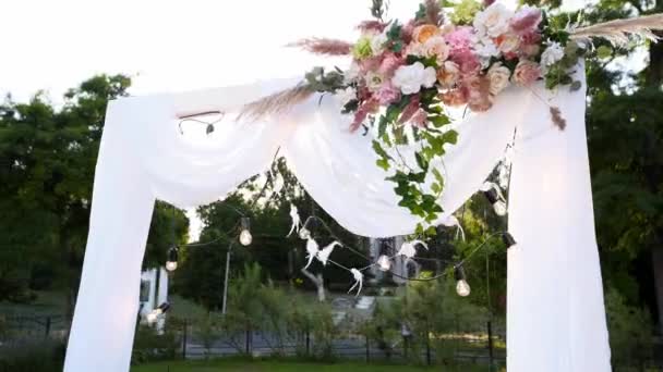 Bröllopsbåge dekorerad med blommor på solig sommardag. Festlig blomma dekoration. Rosor och fjäder gräs med lökkrans på vit textil ram. — Stockvideo