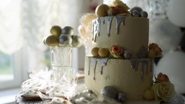 Wedding cake with roses, white glaze and silver topping on candy bar. Cakepops, biscuits and cookies on birthday party served table. Balloons on background. — Stock Video