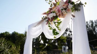 Düğün kemeri, güneşli yaz gününde çiçeklerle süslenmiş. Şenlikli çiçek süslemesi. Güller ve tüy çimleri beyaz tekstil çerçevesinde ampul çelengi ile.