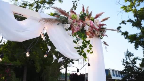 Łuk ślubny ozdobiony kwiatami w słoneczny letni dzień. Świąteczna dekoracja kwiatowa. Róże i trawa piórkowa z girlandą na białej ramie włókienniczej. — Wideo stockowe