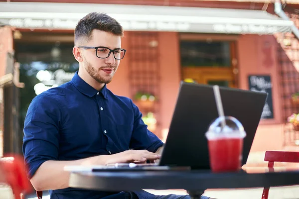 Young attractive hipster freelancer working remotely on laptop at outdoor cafe. Businessman browsing internet at coffeshop. Blogger posts to social media on travel using 5g internet. Telecommuting. — Stock Photo, Image
