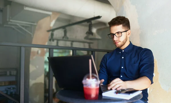 Young attractive hipster freelancer working remotely on laptop in loft cafe. Businessman browsing internet at coffeshop. Blogger posts to social media on travel using 5g internet. Telecommuting theme. — Stock Photo, Image