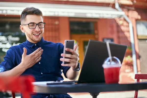 Smiling freelancer with smartphone telecommuting via online video chat. Distracted from work businessman holding mobile phone. Man in glasses with laptop on table in outdoor cafe. Multitasking theme. — Stock Photo, Image