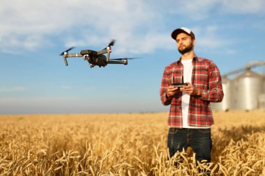 Drone, çiftçinin önünde elinde uzaktan kumandayla tahıl asansörünün yanında asılı duruyor. Çeyrek helikopter pilotun yanında uçuyor. Agronomist buğday tarlasında hava fotoğrafları ve videolar çekiyor.