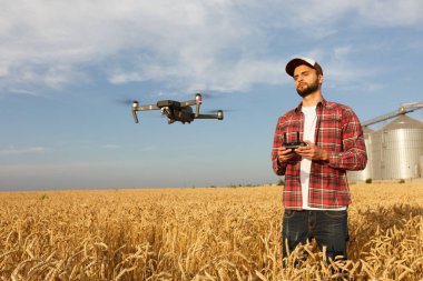 Drone, çiftçinin önünde elinde uzaktan kumandayla tahıl asansörünün yanında asılı duruyor. Çeyrek helikopter pilotun yanında uçuyor. Agronomist buğday tarlasında hava fotoğrafları ve videolar çekiyor.