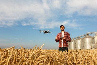 Drone, çiftçinin önünde elinde uzaktan kumandayla tahıl asansörünün yanında asılı duruyor. Çeyrek helikopter pilotun yanında uçuyor. Agronomist buğday tarlasında hava fotoğrafları ve videolar çekiyor.