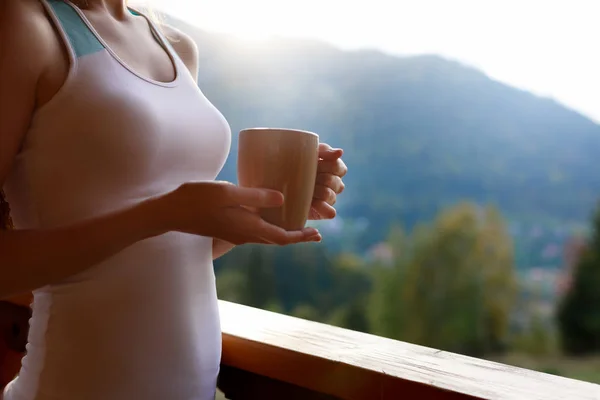 Тонка біла жінка тримає чашку чаю в руках на гірському курорті. Спортивна дівчина з чашкою гарячої кави на дерев'яному балконі заміського будинку. Ліс і гори на фоні. Концепція дієти . — стокове фото