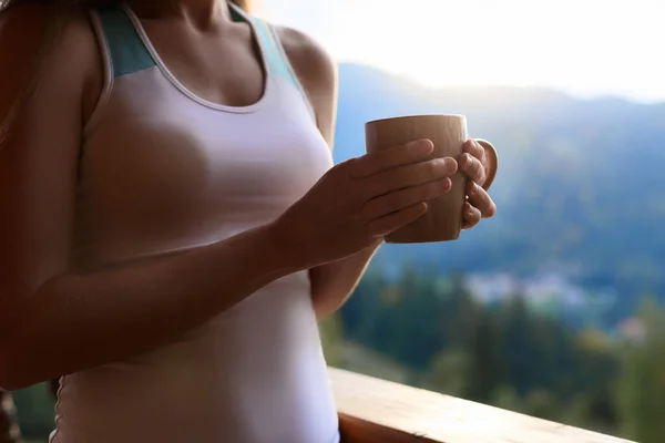 Тонка біла жінка тримає чашку чаю в руках на гірському курорті. Спортивна дівчина з чашкою гарячої кави на дерев'яному балконі заміського будинку. Ліс і гори на фоні. Концепція дієти . — стокове фото