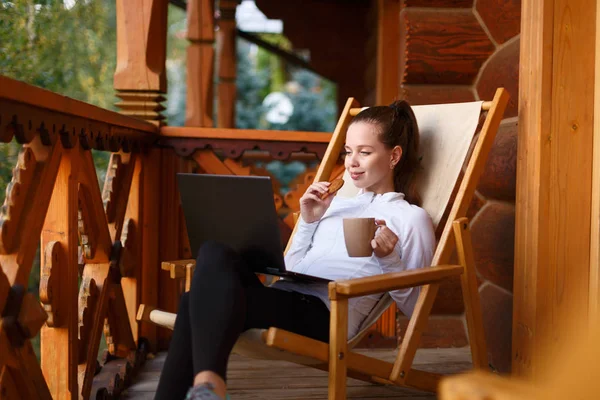 Young attractive business woman is working on the laptop at mountain resort with a cup of tea and cookie. Female relaxes on deck chair with notebook and coffee. — Stock Photo, Image