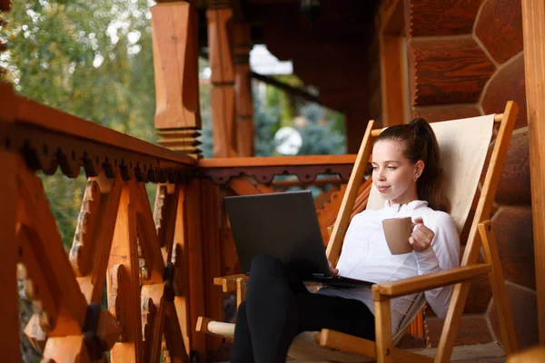 Young attractive business woman is working on the laptop at mountain resort with a cup of tea and cookie. Female relaxes on deck chair with notebook and coffee. — Stock Photo, Image