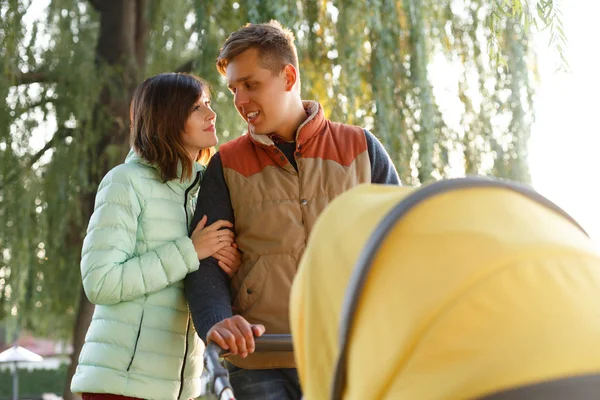 Een jonge liefhebbende familie loopt langs het meer met een wandelwagen. Lachende ouders paren met kinderwagen in het najaarspark. Liefde, ouderschap, familie, seizoen en mensen concept. — Stockfoto
