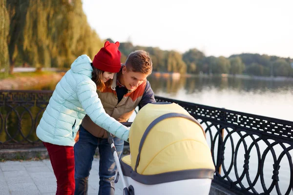 Een jonge liefhebbende familie loopt langs het meer met een wandelwagen. Glimlachende ouders paar met kinderwagen in het najaar park kijken in kariage. Liefde, ouderschap, familie, seizoen en mensen concept. — Stockfoto