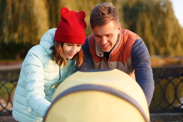 Een jonge liefhebbende familie loopt langs het meer met een wandelwagen. Glimlachende ouders paar met kinderwagen in het najaar park kijken in kariage. Liefde, ouderschap, familie, seizoen en mensen concept. — Stockfoto