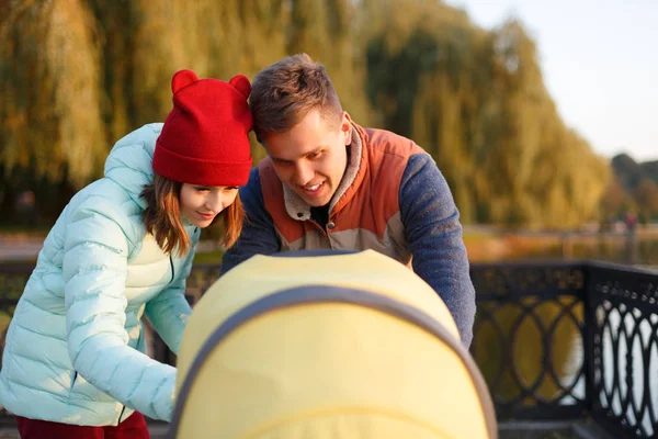 Een jonge liefhebbende familie loopt langs het meer met een wandelwagen. Glimlachende ouders paar met kinderwagen in het najaar park kijken in kariage. Liefde, ouderschap, familie, seizoen en mensen concept. — Stockfoto