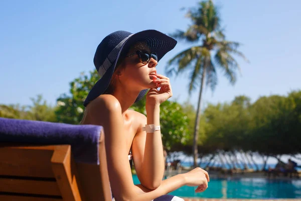 Retrato retocado de mujeres jóvenes en sombrero de paja y playa elegante disfrutando del sol tropical y sentado en una tumbona. Concepto de cuidado de piel de verano. Linda chica de vacaciones en la isla cerca del océano — Foto de Stock