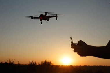 Helikopter arka planda uçarken adam uzaktan kumandayı elleriyle tutuyor. Drone, Suset 'teki pilotun arkasında duruyor. Yüzü yok