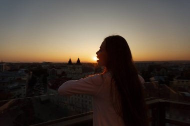 Balkondan yükselen Avrupa sokak mimarisinin panoramik manzarasına hayran olan genç ve güzel bir kadın. Gün batımında sokak manzarasına bakan bir kız. Mutlu dişi güneş yerken eğleniyor..