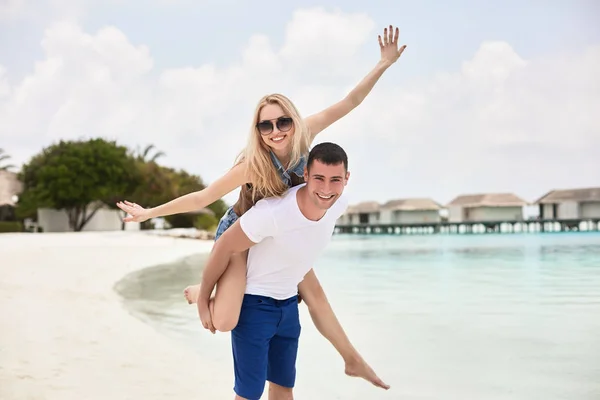 Retrato de sorrir homem carregando mulher em suas costas ao longo da costa do mar. cara dando piggyback passeio para namorada pelo oceano. Casal jovem se divertindo na praia resort tropical. Conceito lua de mel. — Fotografia de Stock