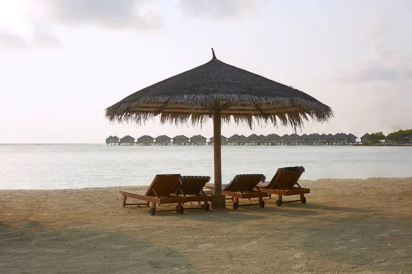 Beach deck chairs under straw umbrellas. Indian ocean coastline on Maldives island. White sandy beach and calm sea. Travel and vacation concept. — Stock Photo, Image