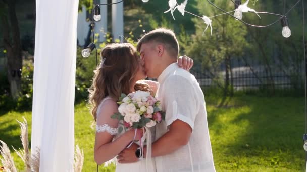 Gelin ve damat dışarıda düğün kemeri altında öpüşüyorlar. Arkadaşlar yeni evlilerin üstünde konfeti vurur. Yaz düğününde çift. — Stok video