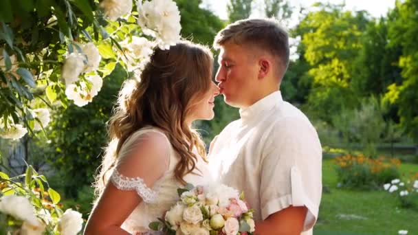 La novia y el novio sonríen y se besan cerca del exuberante arbusto de rosas blancas. Los recién casados se embellecen y miran amorosamente a los ojos del otro. Hermosa pareja en vestidos de novia blancos. Escena retroiluminada . — Vídeo de stock