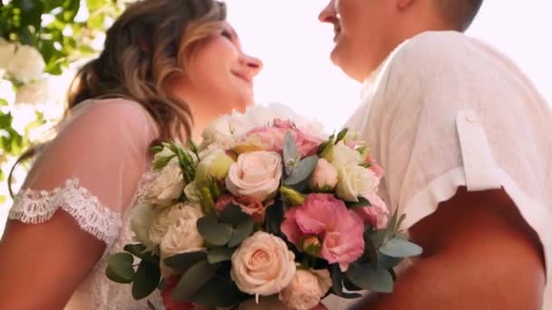 Mariés et mariés sourient et s'embrassent près luxuriante rosiers blancs buisson. Les jeunes mariés s'embrassent et se regardent avec amour les yeux. Beau couple en robes de mariée blanches. Scène rétroéclairée . — Video