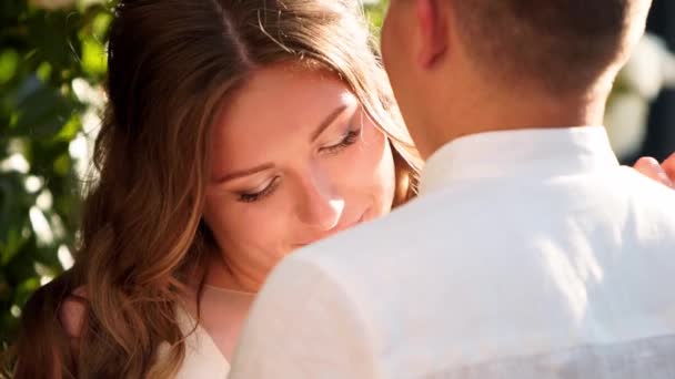 Bride and groom are smiling and kissing near lush white roses bush. Newlyweds embace and look lovingly to each others eyes. Beautiful couple in white wedding dresses. Backlit scene. — Stock Video