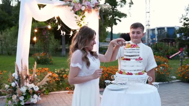 新婚旅行は屋外の装飾されたアーチの近くの暖かい夏の日没の結婚式のケーキを切った。花嫁と新郎は笑顔と1つのナイフで手と一緒に結婚式のケーキをカット。スローモーション. — ストック動画