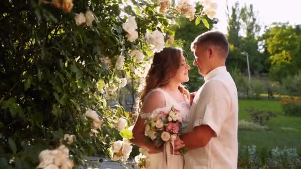 La novia y el novio sonríen y se besan cerca del exuberante arbusto de rosas blancas. Los recién casados se embellecen y miran amorosamente a los ojos del otro. Hermosa pareja en vestidos de novia blancos. Escena retroiluminada . — Vídeo de stock