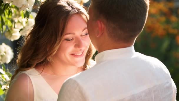 La novia y el novio sonríen y se besan cerca del exuberante arbusto de rosas blancas. Los recién casados se embellecen y miran amorosamente a los ojos del otro. Hermosa pareja en vestidos de novia blancos. Escena retroiluminada . — Vídeo de stock