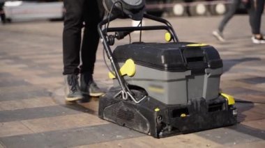 Kapıcı elle itilen elektrikli süpürge arabası ve şehir kaldırımlarını temizliyor. Kapısı ıslak, sıcak, kaplamalı levhalar açık havada. Yol temizleme makinesi detayları. Karantina için hijyenik önlemler.