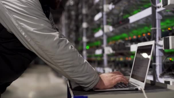 Data center diagnostics technician works and types on laptop. Mining equipment service in server room. Administrator repairs computing cluster with network hardware lights on background. Slider shot. — Stock Video