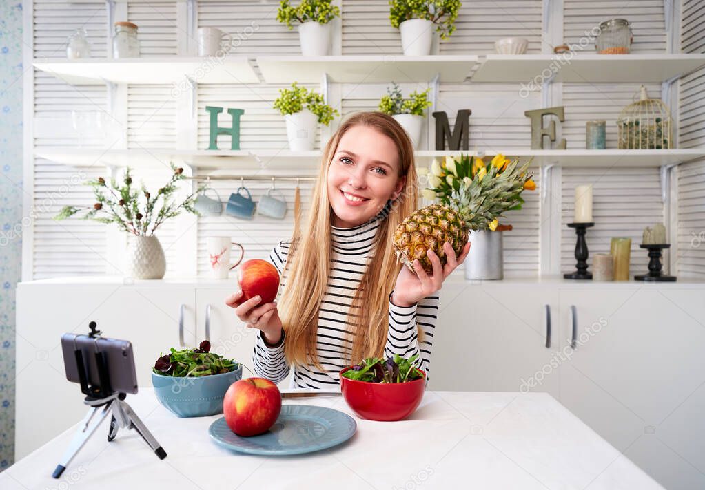 Food blogger cooking fresh vegan salad of fruits in kitchen studio, filming tutorial on camera for video channel. Female influencer holds apple, pineapple and talks about healthy eating. Fructorianism